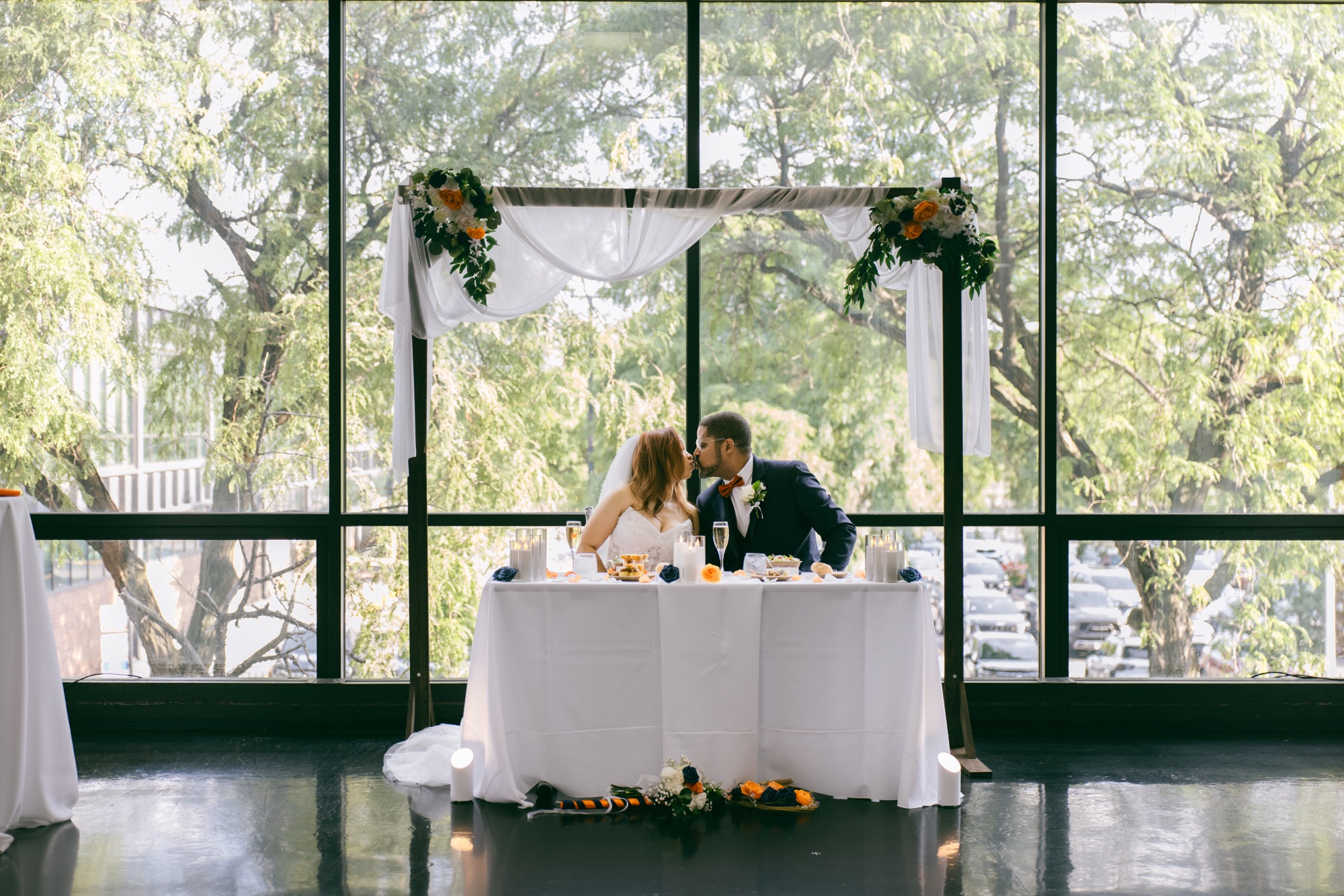 Newlyweds kissing at a sweetheart table before tall windows